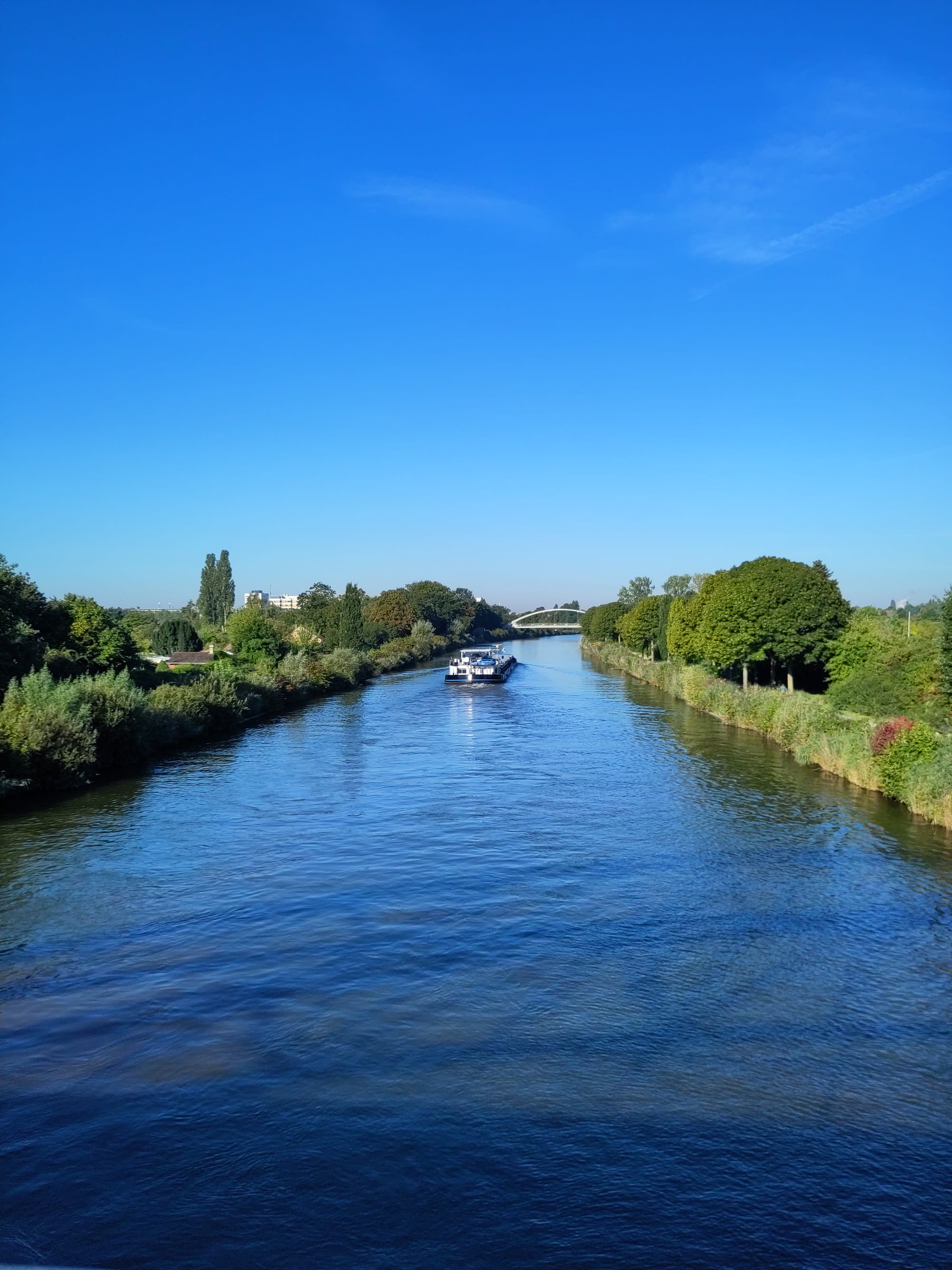 Mittellandkanal mit blauem Wasser. Auf dem Kanal ein Transportschiff, welches sich vom Betrachtenden entfernt. An den Rändern des Kanals grüne Büsche und Bäume. 