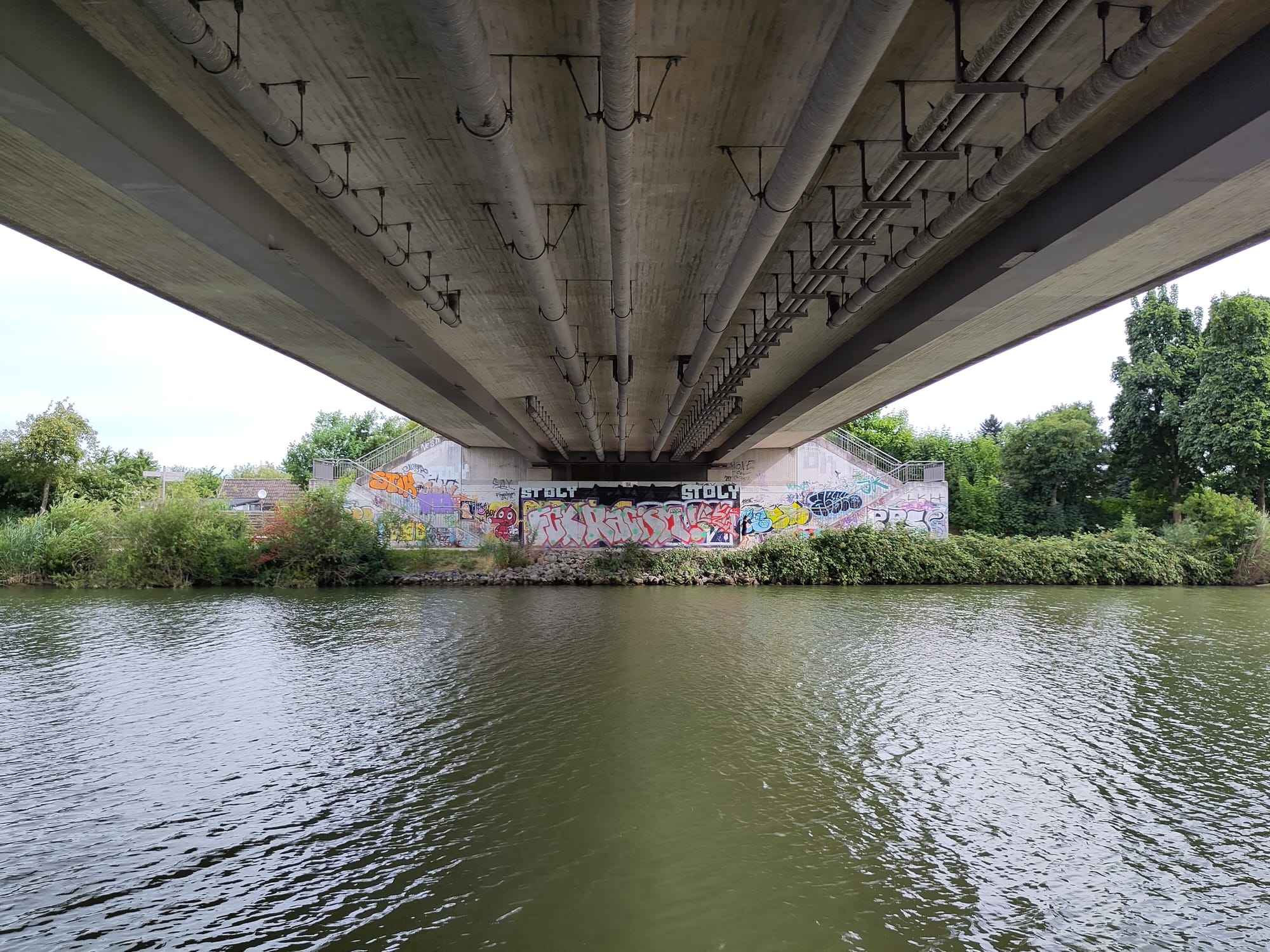 Blick auf den Mittellandkanal von unterhalb einer Brücke. Am Brückpfeiler der gegenüber liegenden Seite Graffiti. Unter der Brückefläche hängen Rohre unterschiedlichen Durchmessers.