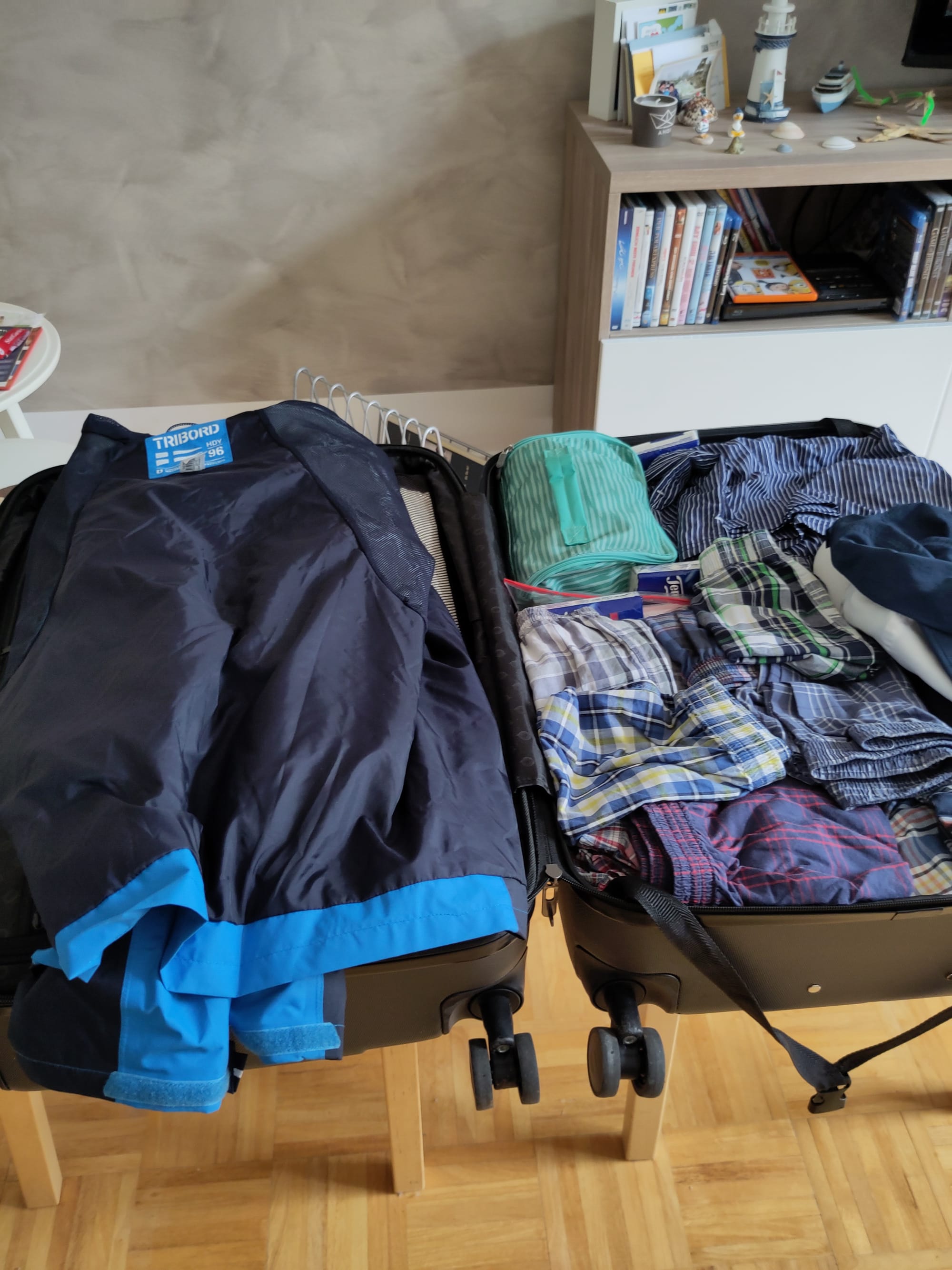 Ein geöffneter Koffer, der auf zwei Stühlen liegt: die linke Seite wird von einer blauen Regenjacke bedeckt, auf der rechten Seite Unterwäsche und einem Kulturbeutel.