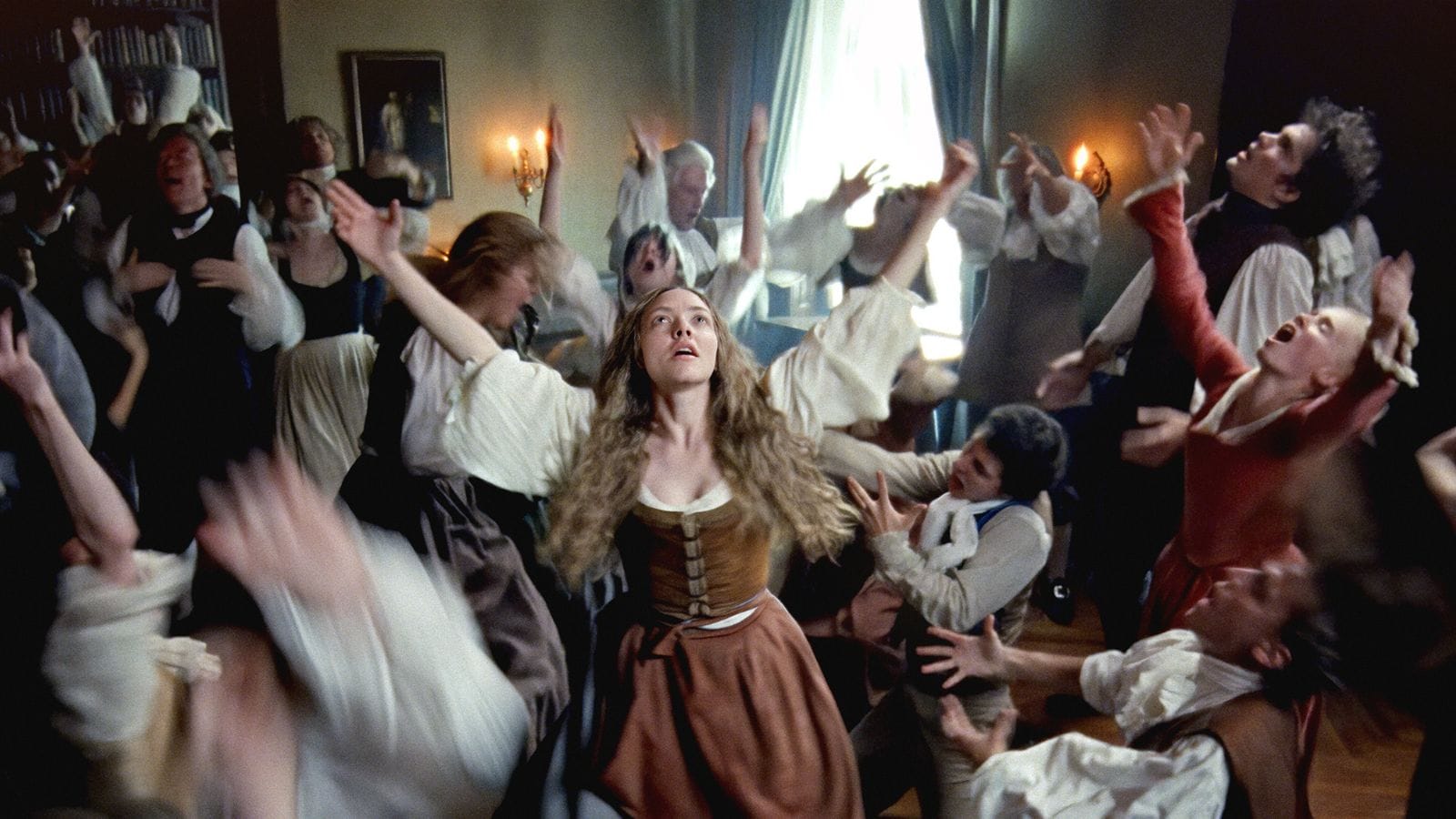 Still image from the 2025 film Testament of Ann Lee: Ann Lee (Amanda Seyfried) stands with her arms wide in the middle of a dancing crowd in 18th century period costumes.