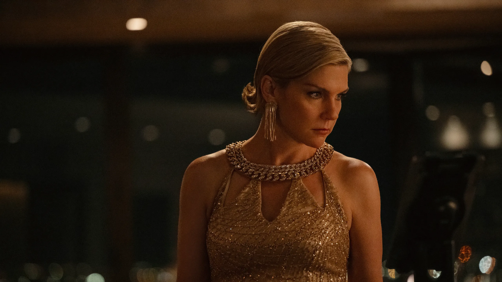 Still from the TV show Pluribus: Carol (Rhea Seehorn) stands alone in a room wearing a fancy gold dress and earrings