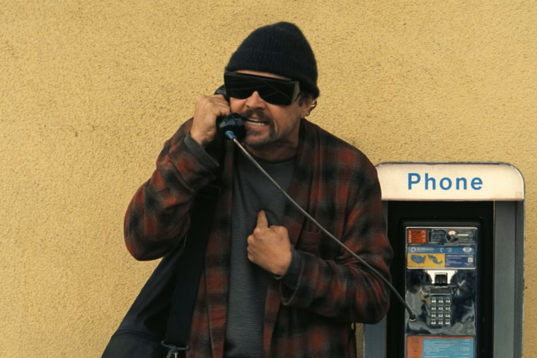 Still image from One Battle After Another: Bob (Leonardo DiCaprio) stands angrily at a payphone against a yellow concrete wall