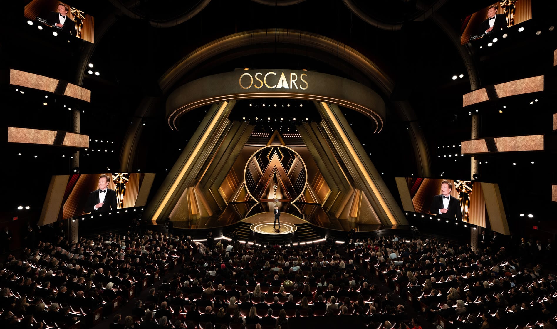 A wide shot of the 2025 Academy Awards: a gold stage with Conan O'Brien presenting in front of rows of attendees.