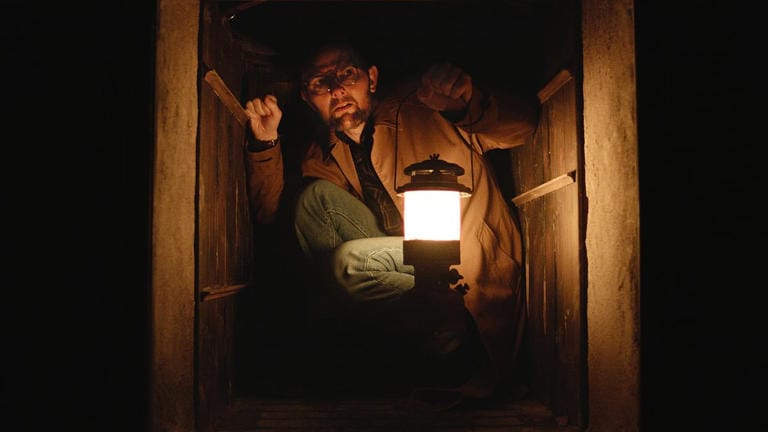 Still from the film Hokum: Ohm Brauman (Adam Scott) is crouched in a dumbwaiter with a lantern