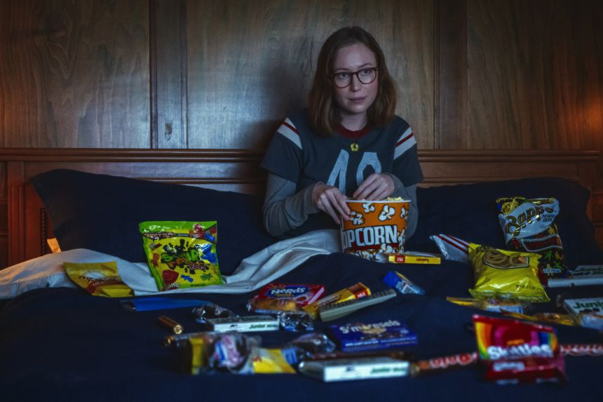 Still from Teenage Sex and Death at Camp Miasma: Kris (Hannah Einbinder) sits in bed eating popcorn, surrounded by candy